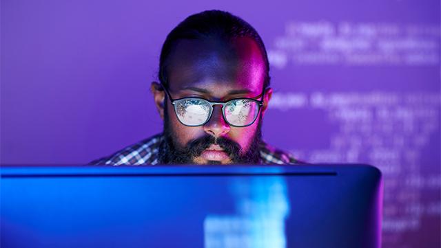 A man behind a laptop with the laptop screen illuminating his face.